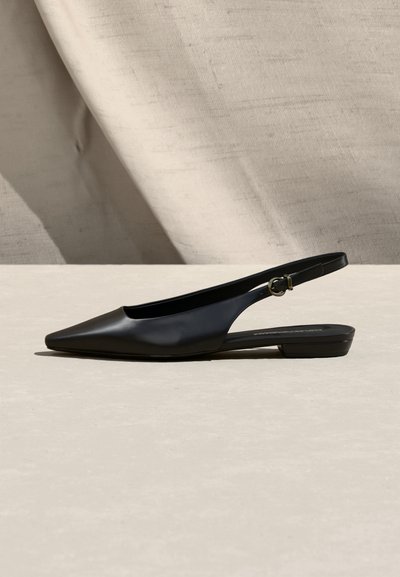 Μαύρο δερμάτινο γυναικείο παπούτσι με αιχμηρή μύτη, φλατ τακούνι και λουράκι πίσω με ρυθμιζόμενη αγκράφα, εμφανιζόμενο από το πλάι πάνω σε μπεζ επιφάνεια.