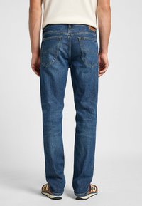 Jeans de mezclilla azul con un corte recto, costuras visibles y un parche de cuero en la parte trasera de la cintura. Tienen cuatro bolsillos.