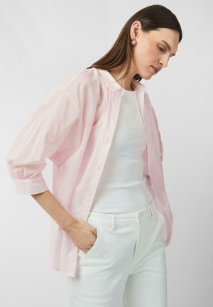 Light pink button-up shirt with a relaxed fit, featuring balloon sleeves. Worn over a white ribbed tank top and white pants, showcasing a minimalist design.