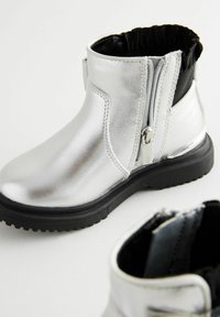 Silberne metallische Knöchelstiefel mit glatter Oberfläche, ausgestattet mit einem schwarzen elastischen Einsatz oben und seitlichem Reißverschluss auf beiden Seiten.