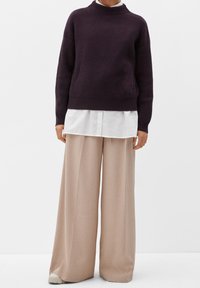 Personne portant un pull côtelé violet foncé sur une chemise blanche, associé à un pantalon beige large et des chaussures claires, se tenant devant un fond blanc.