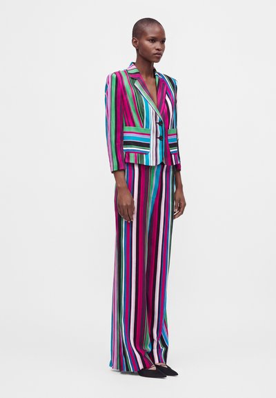 Femme debout portant un costume à rayures verticales colorées avec un blazer ajusté et un pantalon à jambes larges sur un fond blanc uni.