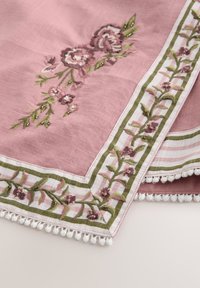 Tela rosa con patrones florales bordados en púrpura y verde, bordeada con ribete blanco y verde, y terminada con flecos de pompones blancos.