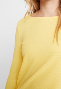 Záběr z blízka na osobu v texturovaném žlutém svetru s dlouhým rukávem, s volnými blond vlasy, zobrazuje krk a horní část trupu.