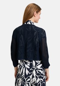 Marineblaue, kurze Strickjacke mit strukturiertem Netzmuster. Sie hat lange Ärmel und einen abgerundeten Saum und ist über einem floral gemusterten Outfit getragen.