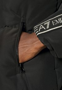 Černá izolovaná bunda s boční kapsou na zip; texturovaná tkanina; stříbrně akcentovaný pás s brandingem "EA7" na rukávu.