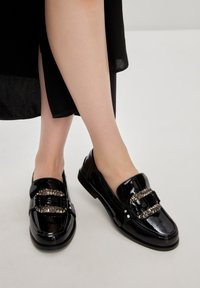 Mocassini in pelle verniciata nera con doppi accenti metallici a catena e suola bassa e piatta. Finitura liscia con punta rotonda e design minimalista.