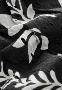 Tissu noir avec des motifs de feuilles blanches, présentant des découpes et une finition texturée. Le matériau semble doux et drape de manière fluide.