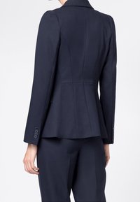 Námořnický modrý oblekový blazer s dlouhými rukávy, strukturovaným střihem a jemnými švy. Obsahuje knoflíkové manžety a hladkou texturu látky.