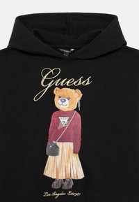 Hoodie noir présentant une illustration d'un ours portant une chemise rouge avec un logo "Guess", une jupe plissée, des chaussures noires et un petit sac à main.