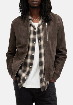 Brown suede bomber jacket with a zipper, layered over a plaid flannel shirt and white ribbed t-shirt, paired with black jeans.