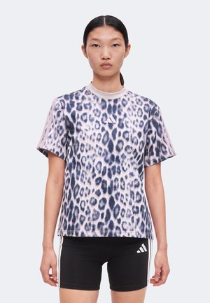 Mladý človek s rovnými čiernymi vlasmi zviazanými vzadu, ktorý má na sebe tričko s krátkym rukávom so vzorom leopardíka a čierne krátke nohavice Adidas s bielymi pruhmi.