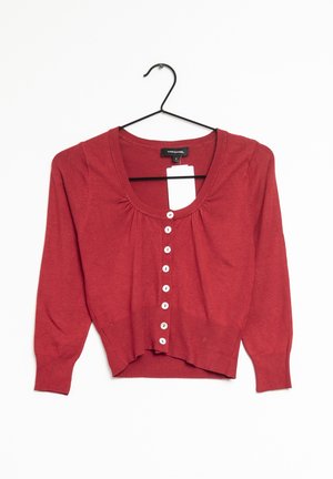 Cardigan court rouge avec manches longues et boutons blancs, accroché sur un cintre noir contre un fond blanc.