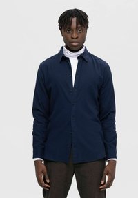Mørkeblå langærmet skjorte med knaplukning og klassisk krave samt sorte knapper, båret over en hvid turtleneck, lavet af bomuldsstof med en glat tekstur.