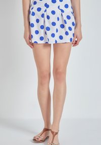 Pantalones cortos blancos con lunares azules, con pliegues en la cintura. El material parece ligero. Combinados con sandalias marrones.