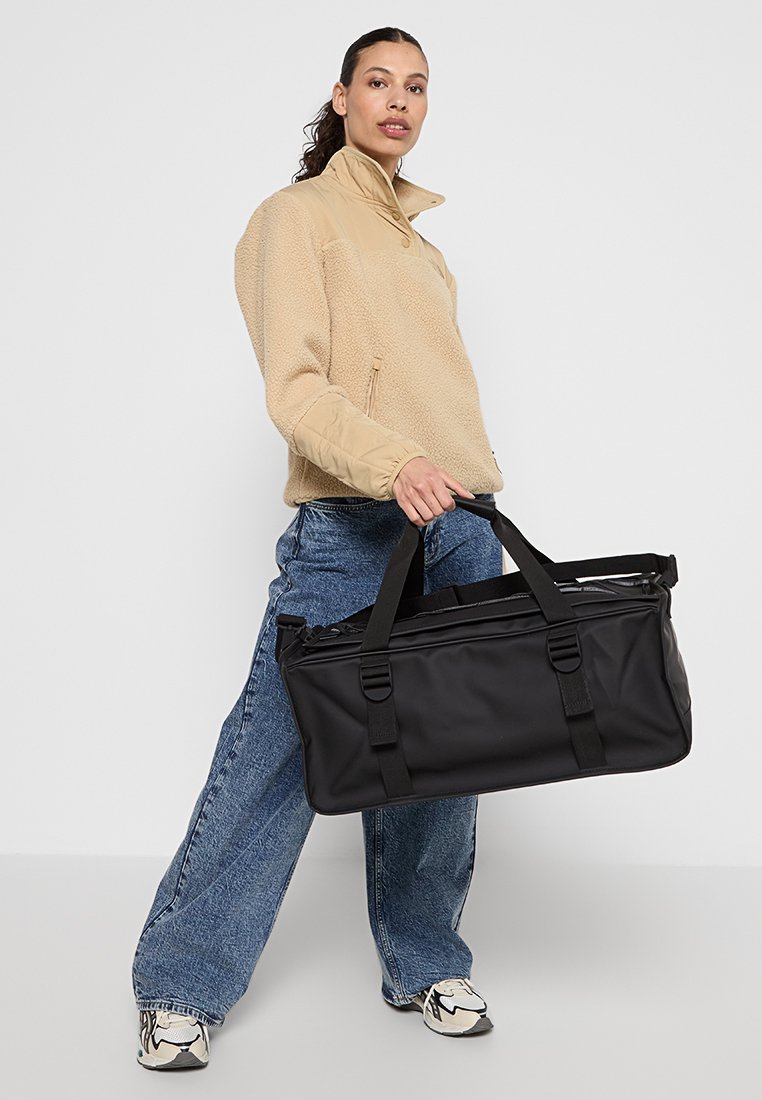 Sac de sport noir avec deux poignées et une fermeture éclair. Modèle portant une veste en polaire beige et un jean en denim ample. Fond neutre.
