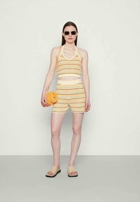 Top cropped de malha riscada e calções em tons de amarelo e laranja, combinado com uma pequena mala laranja. O modelo usa óculos de sol pretos e chinelos em tom de pele.