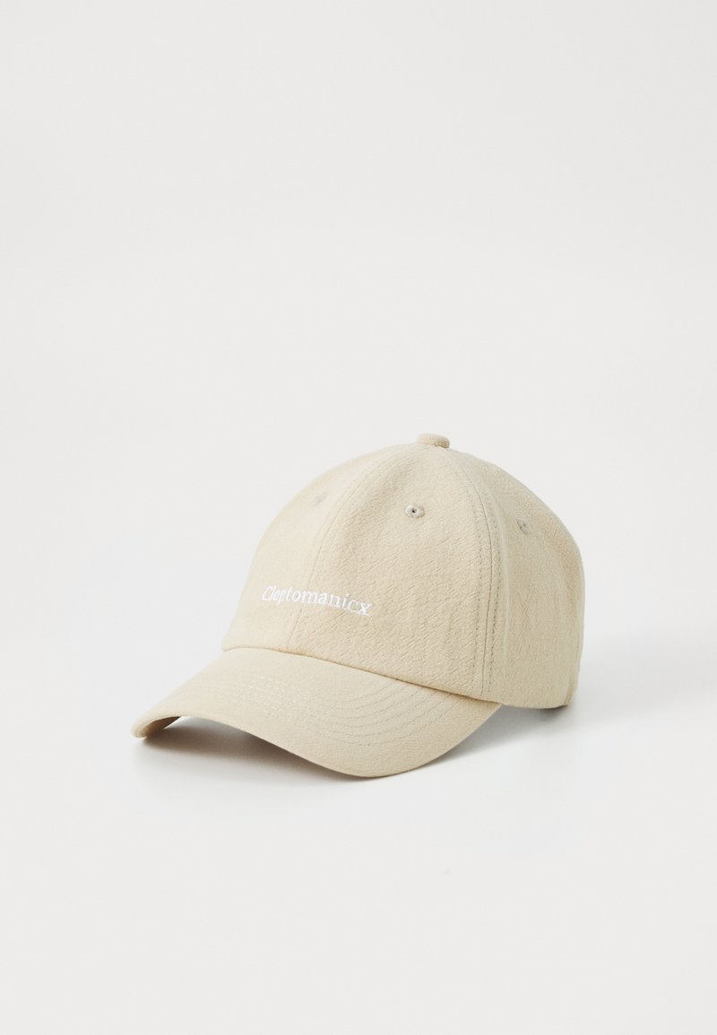Gorra de béisbol beige hecha de tela texturizada. Presenta un ala curvada, seis paneles y el logotipo bordado de "Cleptomanicx" en blanco.