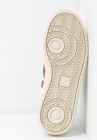 Suela de zapatilla con una superficie de goma beige texturizada, que presenta patrones circulares y en zigzag, con un acento burdeos en el costado y detalles del logo.
