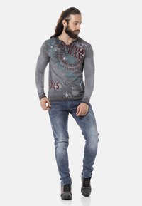 Langärmliges graues Shirt mit grafischen Drucken in Türkis und Burgunderrot, kombiniert mit hellblauen Denim-Jeans und dunkelgrauen Schnürstiefeln.