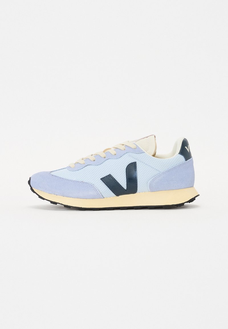 Ljusblå sneakers med mocka- och nätmaterial, krämfärgade snören, mörkblå logotyp och gummisulor. Har en rundad tå och kontrasterande detaljer.