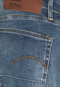 Jeans in denim blu con un'etichetta in pelle contrassegnata "RAW." Presentano cuciture gialle e un piccolo particolare della tasca. Il tessuto ha una texture standard.