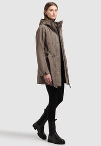 Ženska, oblečena v taupe plašč s kapuco in vezalkami, črne legice ter črne škornje do sredine meč, stoji pred enakomerno svetlo ozadje.