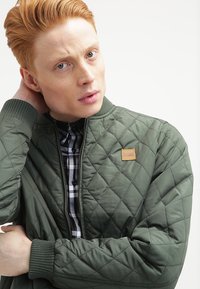 Jachetă bomber verde matlasată, cu manșete ribbate, având un patch cu logo și o cămașă în carouri alb-negru dedesubt. Textură netedă.