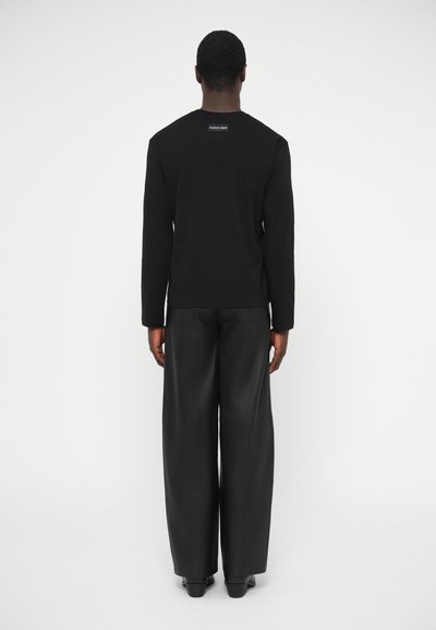 Camisa preta de manga comprida com etiqueta na parte de trás, combinada com calças largas pretas de couro sintético, vista de trás contra um fundo simples.