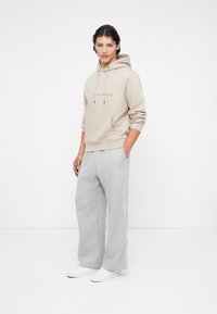 Ung mand iført en beige hættetrøje med frontlomme, lysegrå brede sweatpants med sidestribe og hvide sneakers, stående med hænderne i lommerne.