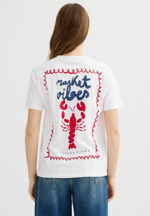 Persona que lleva una camiseta blanca con una langosta roja y el texto "market vibes" en la parte trasera, combinada con vaqueros azules.