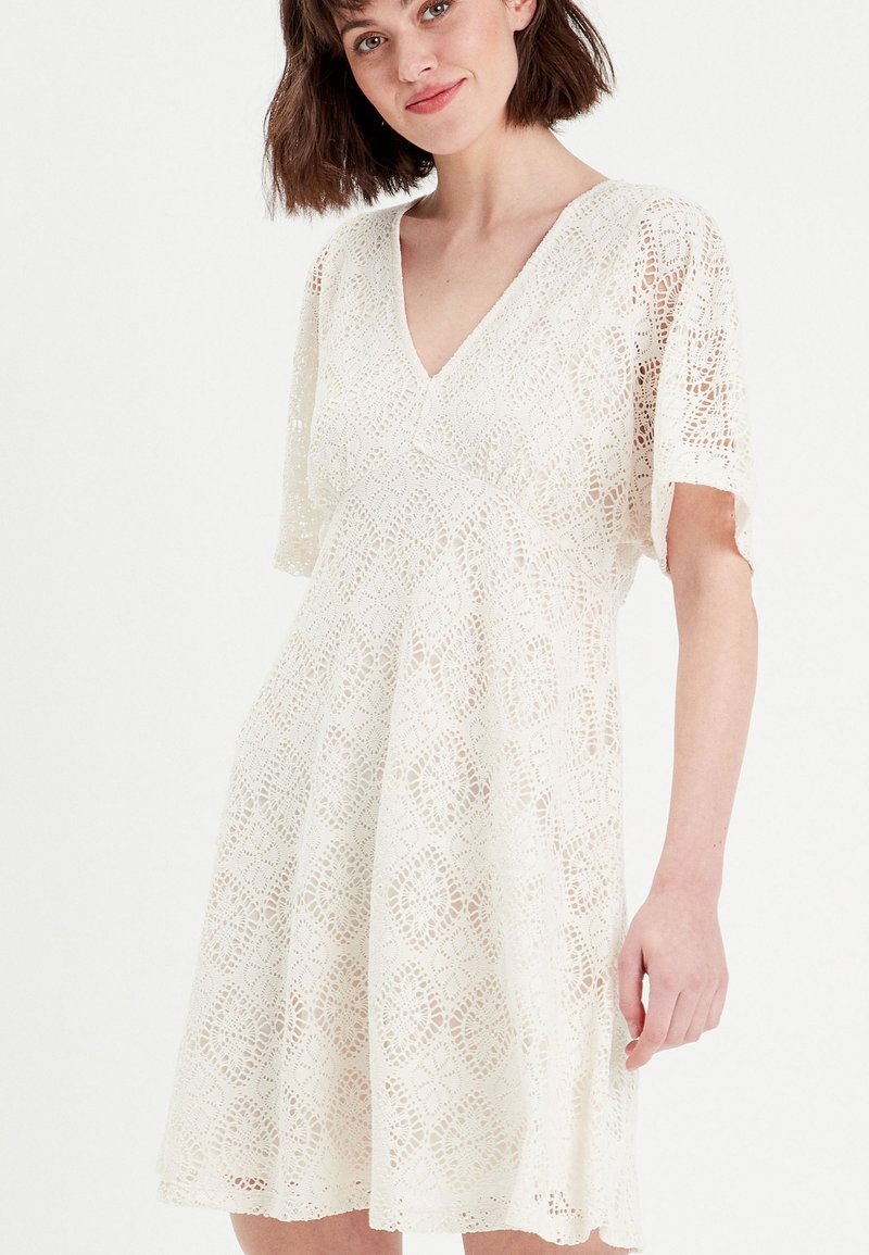 Robe en dentelle de couleur écru avec un décolleté en V et des manches courtes. Présente des motifs floraux complexes, une jupe évasée et une texture délicate.