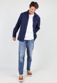 Chemise bleue marine à boutons avec un t-shirt blanc à col rond, associée à un jean bleu clair et des baskets marron. Coupe décontractée, tissu doux et design simple.