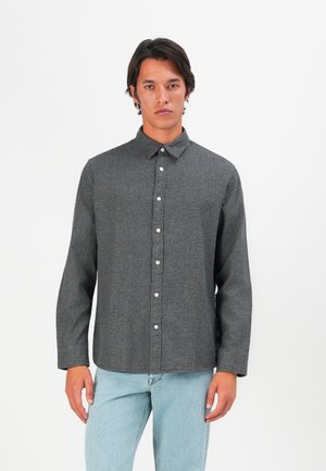 Chemise grise à manches longues avec un col, en tissu texturé. Présente des boutons blancs et un ourlet droit. Portée avec un jean bleu clair.