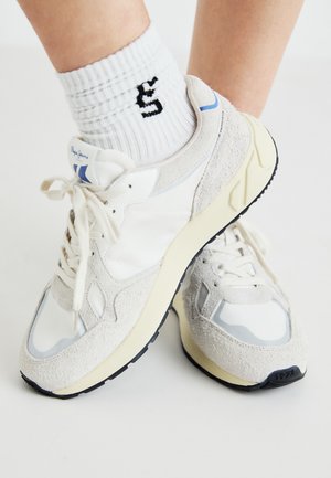 Fehér és világosszürke sportcipők, amelyek velúr és szintetikus anyagokból készültek, kerek orrúak, fűzős kialakítással rendelkeznek, és fekete gumitalppal, valamint logóval vannak ellátva.