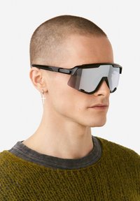 Mustad sportlikud päikeseprillid nurga all disainiga, peegeldavate hallide läätsedega ja paksud külgedega. Kandmas mudel, kellel on lühikesed juuksed ja tekstuuriga rohelise värviga kampsun.