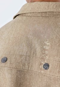 Chaqueta vaquera beige con un acabado texturizado, dos botones de metal negro y costura visible a lo largo de la costura trasera.