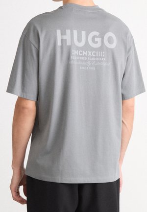 Person iført en grå kortærmet t-shirt med stor "HUGO" og mindre tekst trykt på øverste del af ryggen.