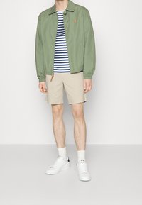 Veste zippée vert clair avec un col, associée à une chemise rayée bleue et blanche, un short beige et des baskets blanches avec des détails de couture.