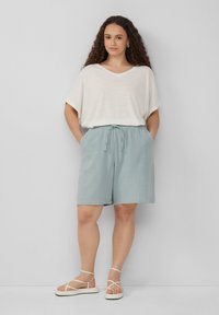 Jasno beżowa koszulka typu V-neck w połączeniu z pastelowymi, niebieskimi szortami do kolan wykonanymi z teksturowanej tkaniny, z regulowanym sznurkiem. Białe sandały na paski.