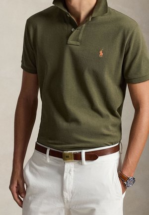 Homme portant un polo vert rentré dans un pantalon blanc avec une ceinture marron et une montre-bracelet au poignet gauche.
