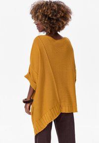 Mosterdgele gebreide poncho met een losse pasvorm, ribbelzoom en een brede halslijn. Heeft een asymmetrische snit en zijdetails.