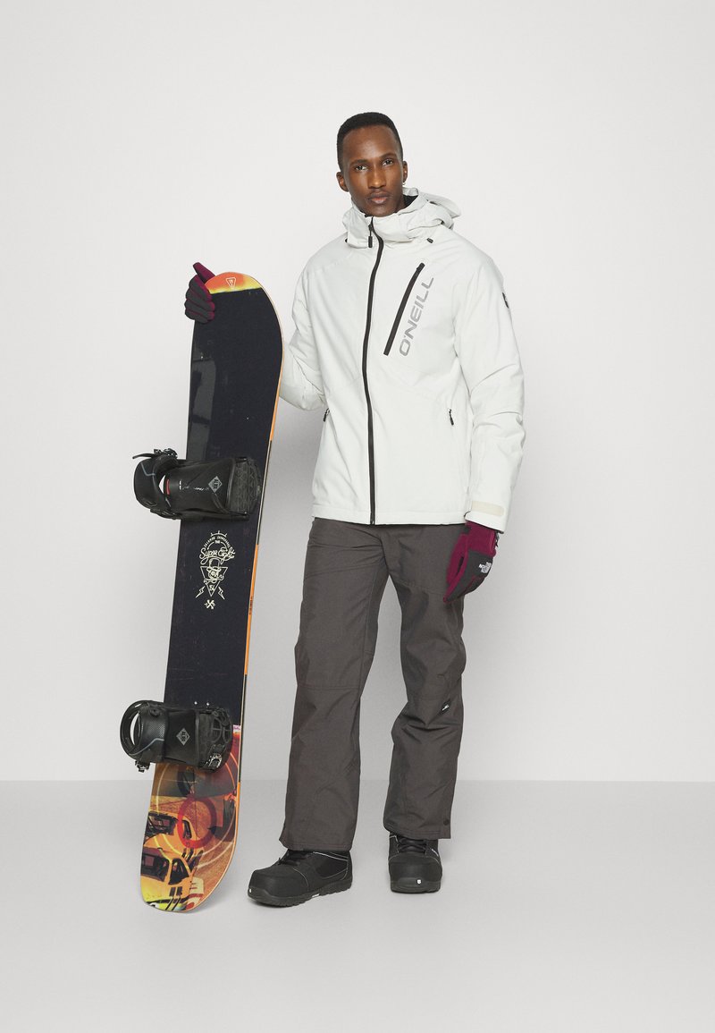 Weißer Jacke mit schwarzem Reißverschluss, dunklen Hosen und schwarzen Schneestiefeln. Hält ein Snowboard mit orangefarbenen und gelben Akzenten sowie Bindungen.