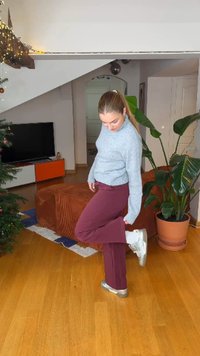 Pull en tricot gris, pantalon bordeaux et baskets argentées. Intérieur avec sol en bois, plante et canapé en arrière-plan.