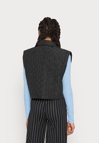 Gilet trapuntato nero con colletto, design senza maniche e motivo a diamante testurizzato, abbinato a una maglietta a maniche lunghe blu chiaro e pantaloni a righe.