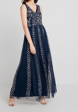 Frau trägt ärmelloses dunkelblaues Abendkleid mit silberner Blumenstickerei und transparentem Überzug, steht auf hellem grauem Hintergrund.
