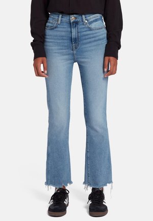 Jean flare bleu clair en denim, avec une finition délavée et un ourlet brut. Porté avec des baskets noires et une chemise sombre.