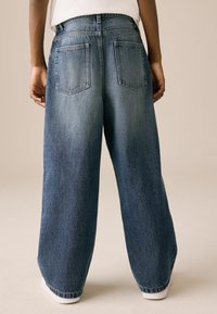 Széles lábú kék denim farmer, tartós anyagból készült, két hátsó zsebbel, kopottas mosással, taperelt szegéllyel és bokáig érő fazonnal.