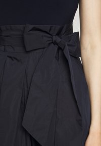 Lähikuva tummansinisestä kankaasta, joka on solmittu suureen nauhaan vyötärölle pleatatuissa hameessa tai mekossa, vierekkäin paljastavan käsivarren kanssa.