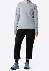 Sudadera de forro polar gris claro con cuello de cremallera, combinada con pantalones negros y zapatos de senderismo azules. Presenta un logo en el pecho y puños elásticos.